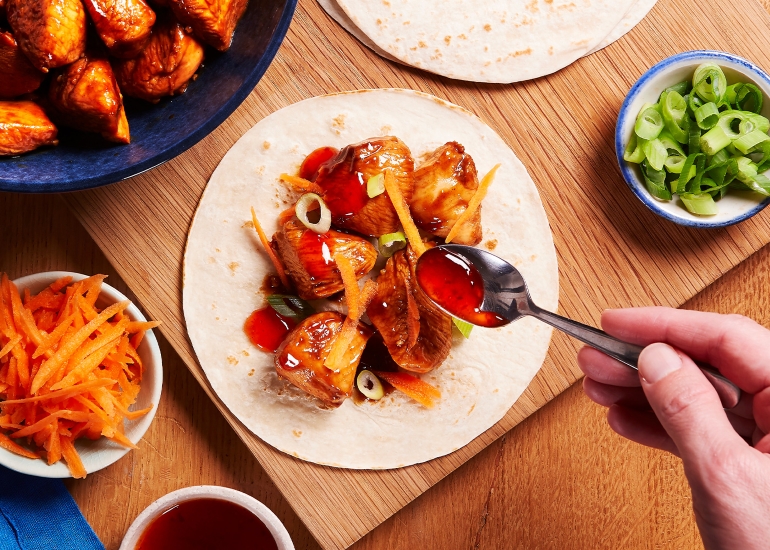 Hand spooning sauce onto tortilla filled with cooked chicken, sliced green onions, and shredded carrots on a wooden board.